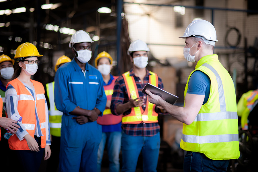 A group of workers in masks gather around a man with an iPad who is conducting a training.